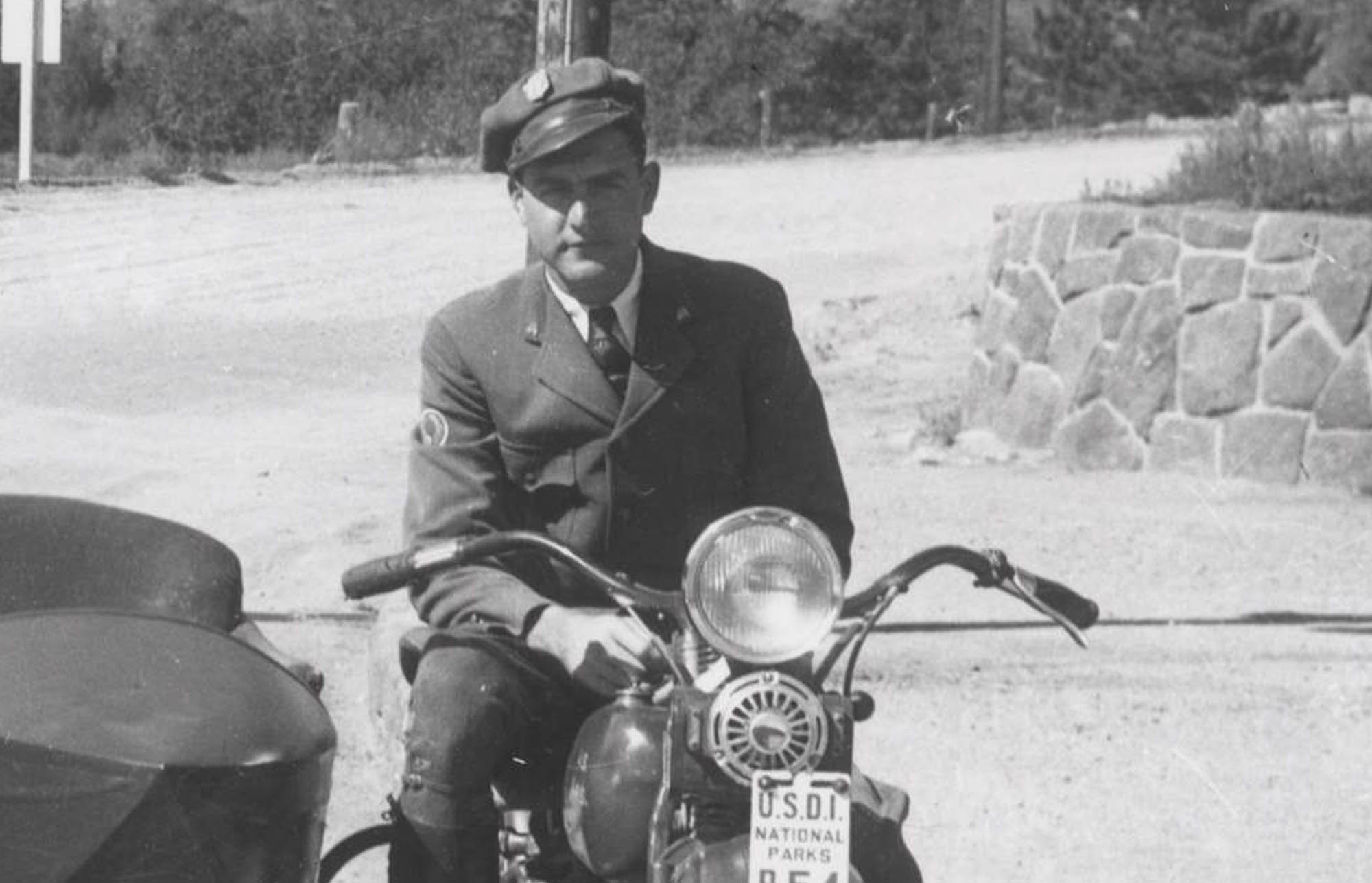 Man in NPS uniform sits astride a motorcycle with sidecar attached beneath a park sign. He wears a shield-shaped badge on his cap.